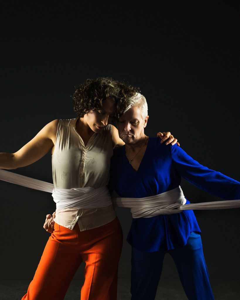 The two dancers, Linda and Helen, stand side-by-side, with their arms around each other’s shoulders, and the edges of their heads are leaning towards each other touching. A caring gesture. Long white fabric is wrapped around each of their waists, connecting them together. They hold the ends of the fabric out to the side with their hands . The fabric symbolises the bond they share as mothers, but could also have reference to the bonds with their children. Linda wears blue clothes, while Helen was a cream top and orange trousers.