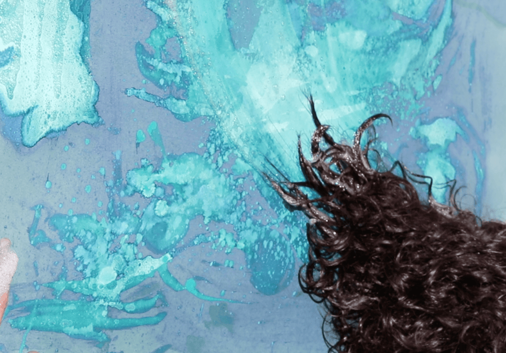 The images shows Helen dragging her hair over the surface of the paper that has been coated in to photosensitive cyanotype chemicals. Her hair if wet and the trails of the water and shapes of the hair leave patterns in the blue chemicals that will expose with the sun.