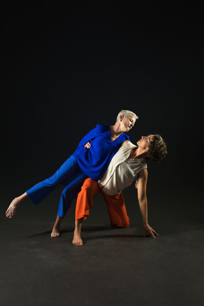 The dancers are in a weight sharing position. Helen, is in a lunge position on the floor, studying herself with one arm as she leans her body sideways towards it. Linda, who is upright, leans onto the side of Helen’s body, with one leg lifted in the air as her body leans the other way over Helen. They look towards each other. The position, that shoes, support and strength, symbolises the strength and support they find in the shared experiences of motherhood.