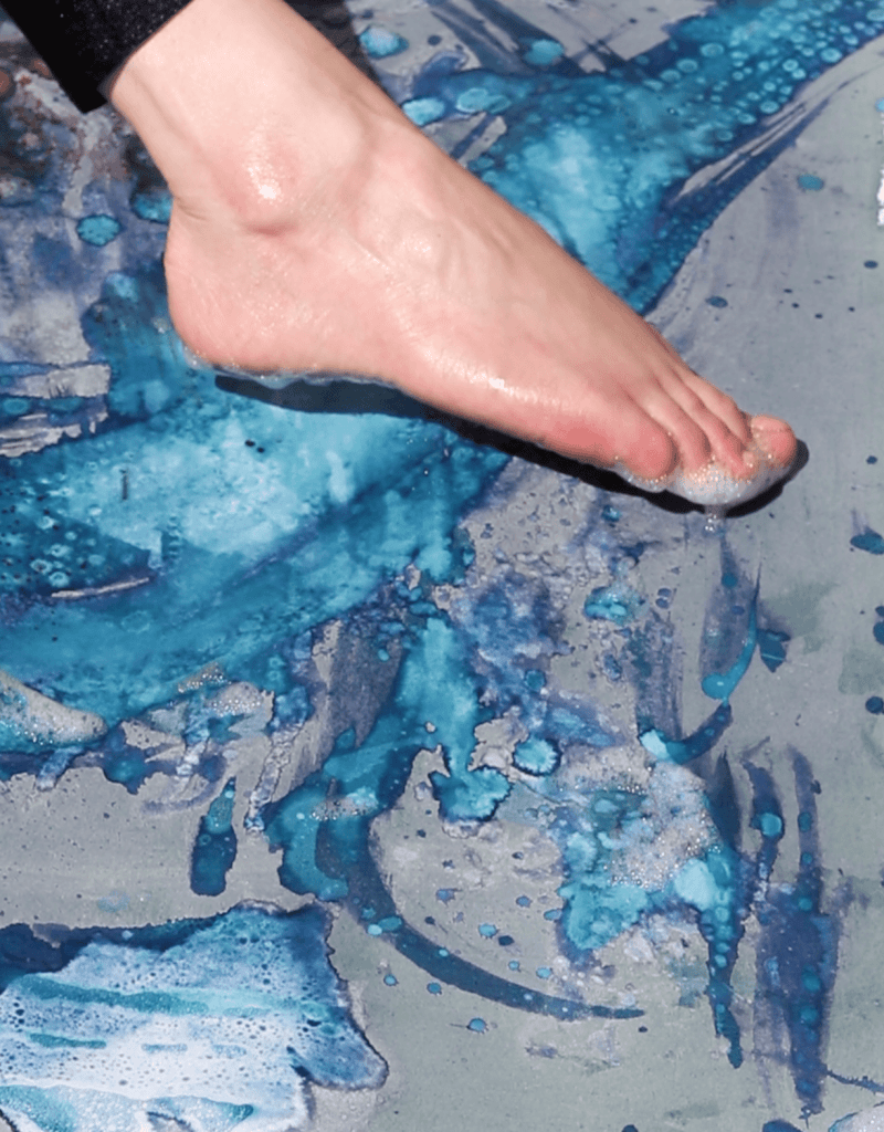 The image shows a close up of Helen’s foot as she sweeps it in gentle movements over the surface of the paper, that has been coated in photosensitive cyanotype chemicals. Her foot leaves patterns in the blue chemicals that will expose with the sun.