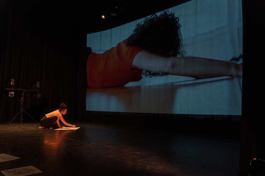 Photo of Helen performing Collecting Moments,, at the Crescent Arts Centre, Belfast, during the Bounce Arts Festival. It shows Helen, wearing a pale orange top and black trousers, crouching down, drawing on a large sheet of paper. At the back of the stage, we see a huge projection screen, showing a video of a previous time Helen danced. She is trying to draw the movements and lines she sees as she watches the giant video projection.