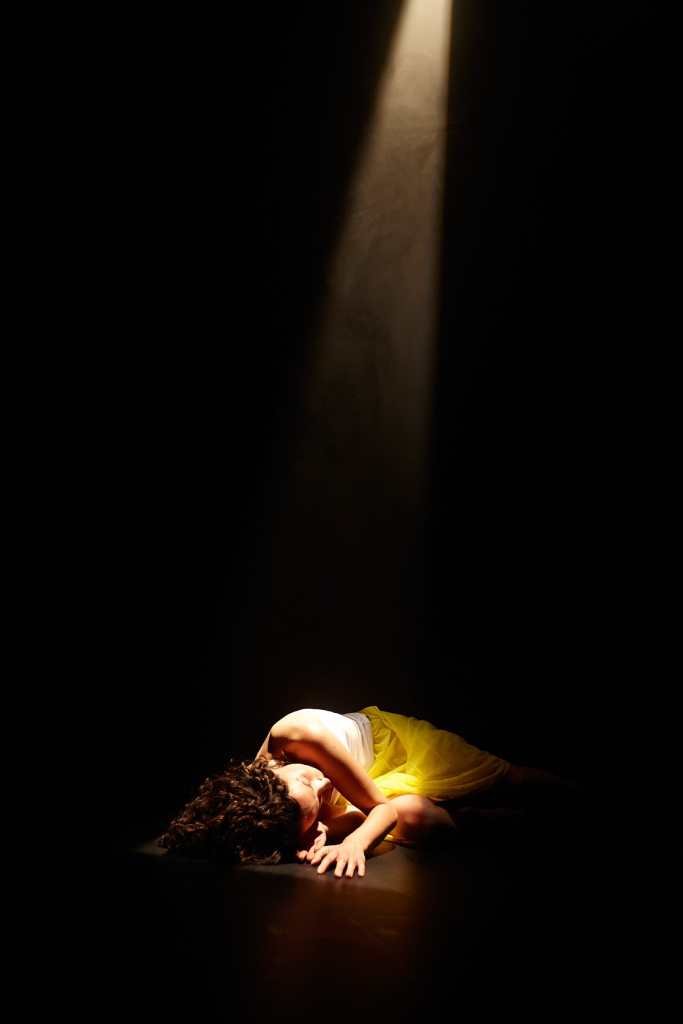 This is a dark image, almost fully black. Helen is lying on the floor on her side. There is a shaft of light coming from above and it lights up part of Helen. She wears a whit top, yellow bottoms and the light glints on the curls in her hair.