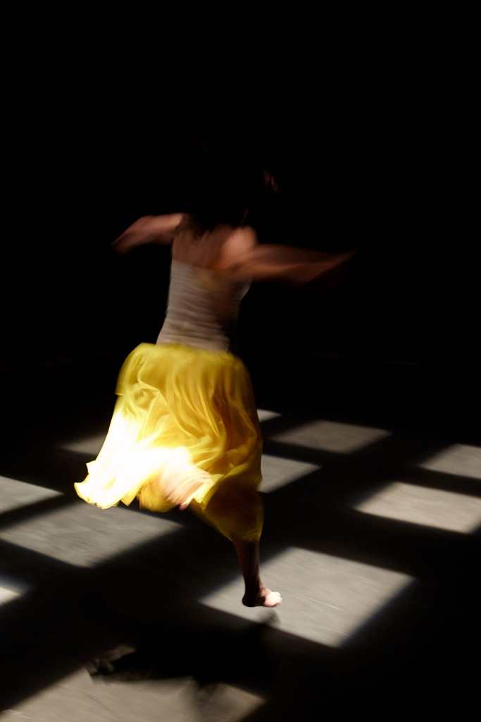 This image captures Helen mid-air while jumping. She jumps form one square of light on the ground to another. The squares of light are in a grid formation. Her Yellow skirt floats behind her.