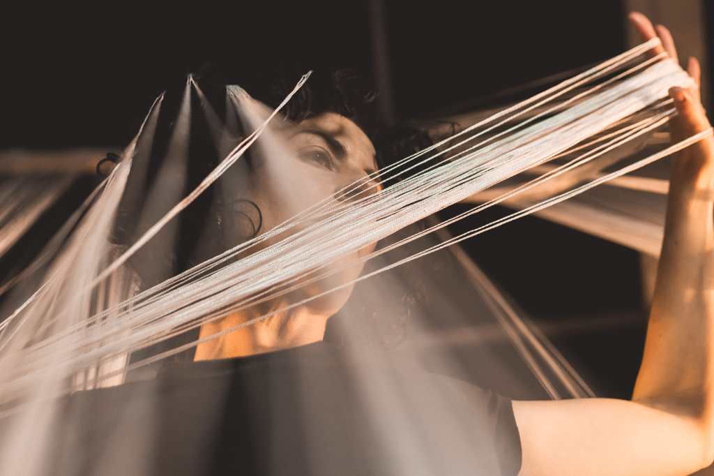 This is quite an abstract image. We see lines of white that are going in many different directions through the image. They almost seem like lines of light. They are actually strings that hang from the ceiling that Helen is moving and playing with. The shot was captured with a slow shutter speed. This is why the lines almost looks like beams of light, and also give a sense of movement and energy. Helen’s face is also seen in the picture, but some of it is hidden behind the strings. Because the image is slightly blurred in places, her black hair almost blends into the background. But we see glimpses of her eyes, her neck, some curls and her hand in amongst the lines of the strings.