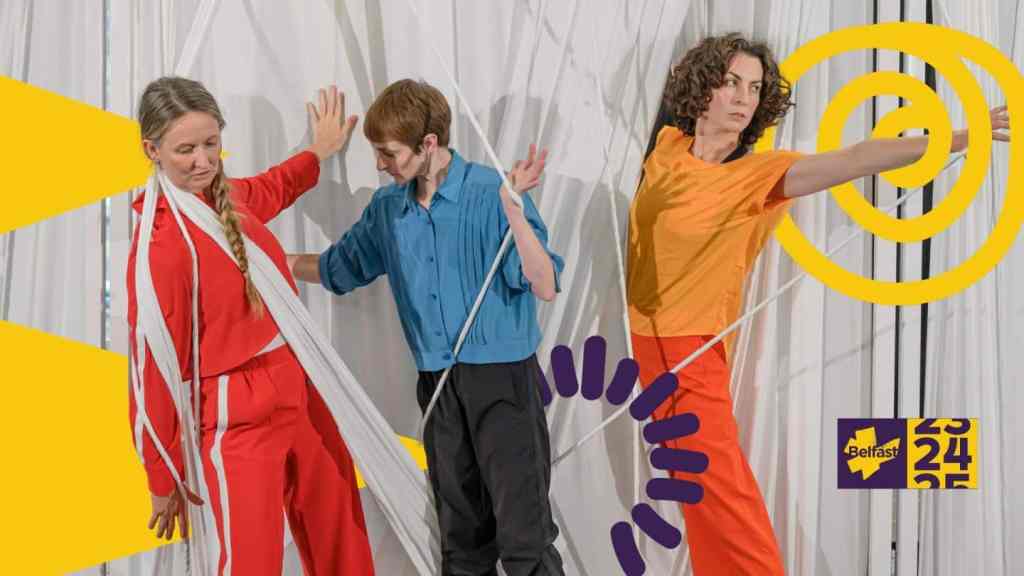 Three dancers wearing bright coloured clothes seem to be emerging from a wall of White fabric. The wall is made of lots of strands of white fabric in different thicknesses. The dancers are wrapped in amongst the wall, with some parts of their bodies behind the wall, and other parts, like an arm or a hand, reaching out towards the camera through the wall. One dancer wears different shades of red clothes. The dancer in the middle wears a bright blue top with a dark bottoms. The dancer on the right wears a bright, yellow top and orange trousers.