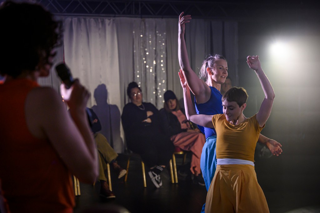 We see two dancers in the midst of a duet. One wears bright yellow clothes and the other wears blue. Their hands reach into the air. On dancer is almost falling back on the other, who supports her weight on the side of her body. It is a duet about one body guiding another in dance and movement. In the foreground we see the third dancer holding a microphone. At this pint she was providing, live, creative audio description.