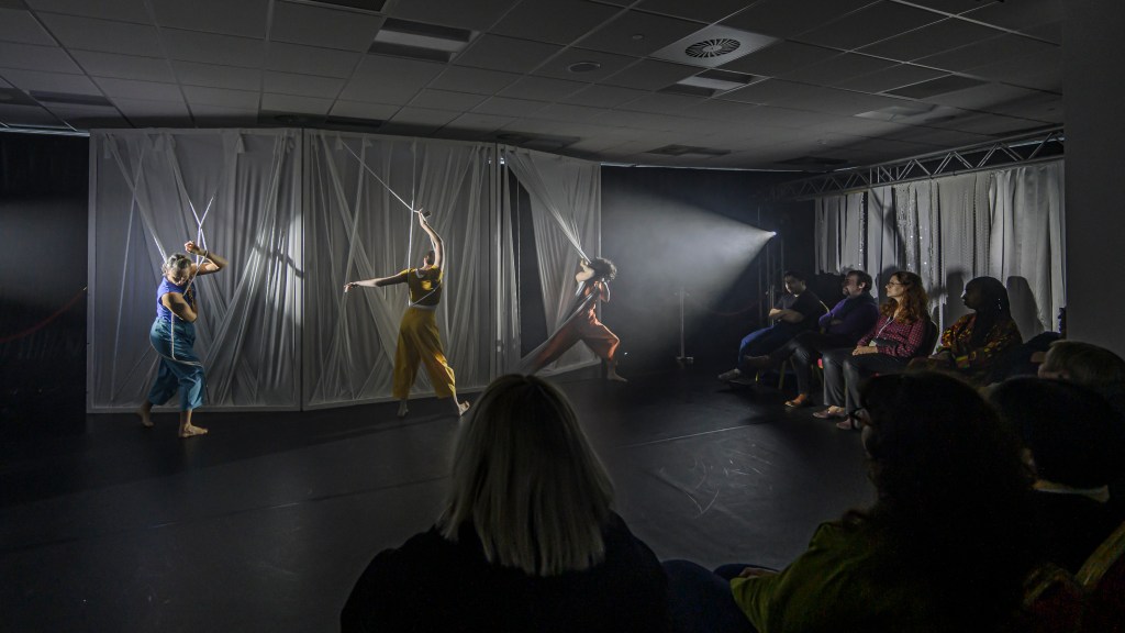 We see all three dancers. They wear bright clothes. One wears yellow clothes, one wears orange clothes and one wears blue clothes. These clothes stand out vividly against a white wall of fabric. The wall is made of strands of stretchy fabric, all in different widths and textures. Some strands are pulled flat and some strands are twisted like a rope. There are three panels of this stretchy white wall. Each dancer stands in front of one of the panels. Each dancer is wrapped in amongst the fabric strands. They are each exploring the textures under their fingertips and wrapping their bodies in amongst the fabric. In this shot, we also see the audience, and we can see that it is a very intimate setting. The audience are seated very close to the dancer.