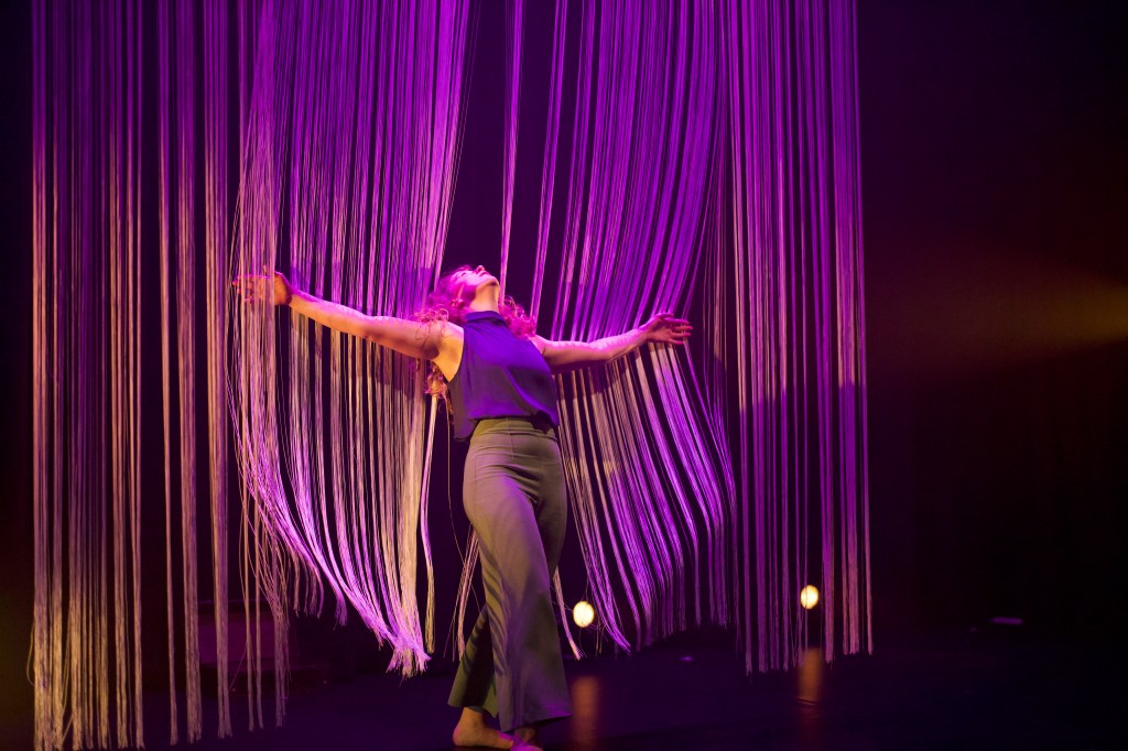 Image of dancer, Rosie, leaning back against a large curtain of hanging string. Her arms are outstretched to the side and as she leans into the string curtain her body makes an imprint as the strings hang around her.