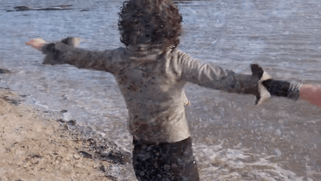 Image shows Helen standing on a beach. We see her from behind. Her arms are stretched out wide to the sides. She look out to sea.