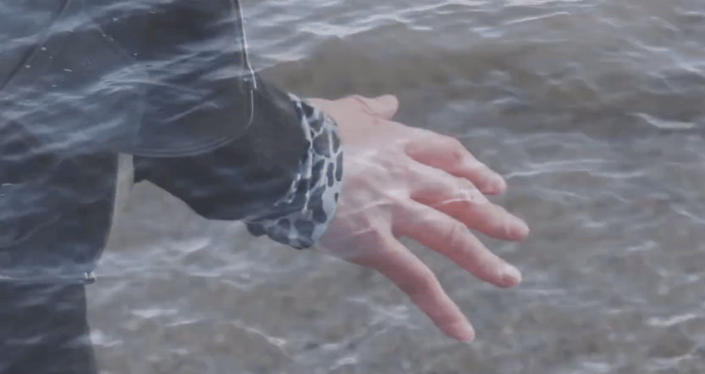 Image shows a close up of the back of Helen's hands. Her fingertips sense the air around her, as she breaths in the sea air. In the background we can see the sea.